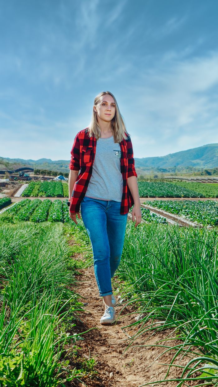 about-01 A blonde woman in casual attire walking through lush green farm fields on a sunny day.